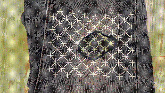 Thick stitches in a komezashi pattern: x's on a diagonal, with stitches running vertically and horizontally between their centers, entering the fabric just a couple millimeters before those points. The pattern covers the knee area of my jeans, from seam to seam across the width of the front panel and six inches high. In the middle is a hole ringed in thick blue thread. Inside the ring, the denim switches from blue to black and the thread changes color from white to green.